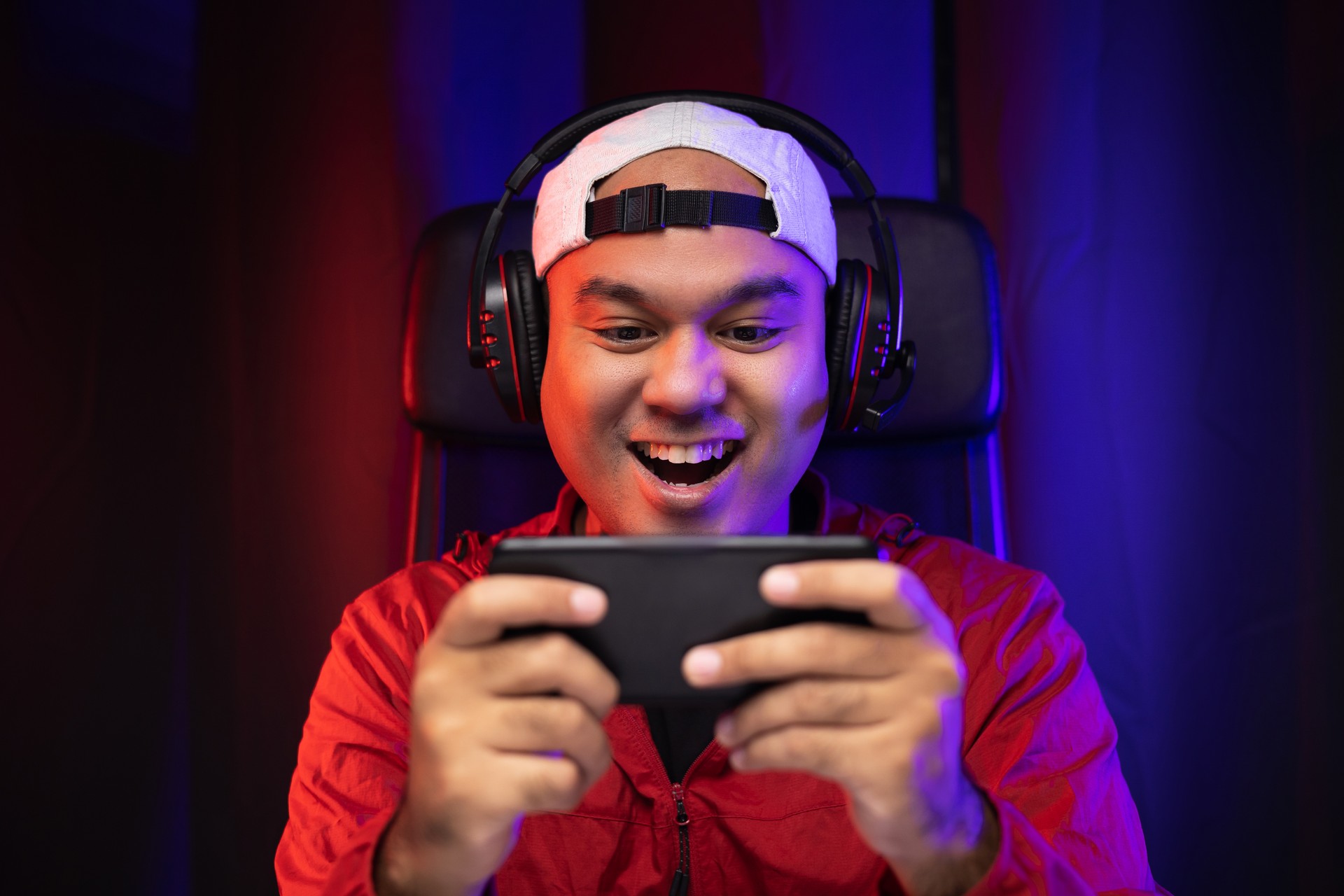 Playing video game on smartphone. Young asian handsome man sitting on chair holding cellphone in living room. Happiness Streamer Indian man wearing headset playing game online in the darkroom.